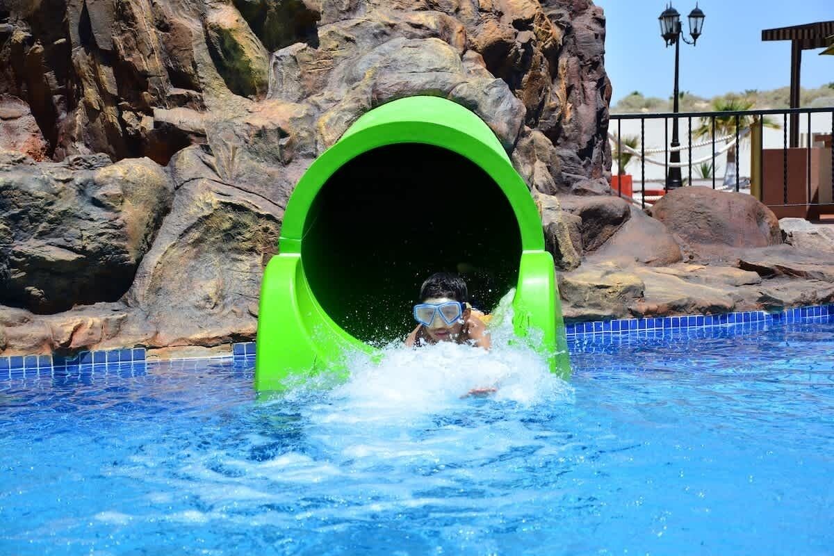 Una persona deslizándose por un tobogán acuático verde hacia una piscina rodeada de rocas.