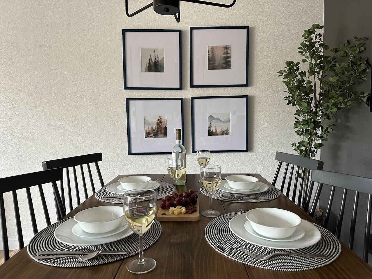 A dining table set with white plates, glasses, and a bottle of white wine, surrounded by black chairs.
