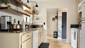 A modern kitchen with white cabinetry, a built-in dishwasher, a stove, and a coffee maker. There is a black mat on the floor and a plant on the countertop.