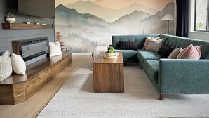 A modern living room with a large flat-screen TV, a wooden coffee table, a teal sofa, and a mountain landscape mural on the wall.
