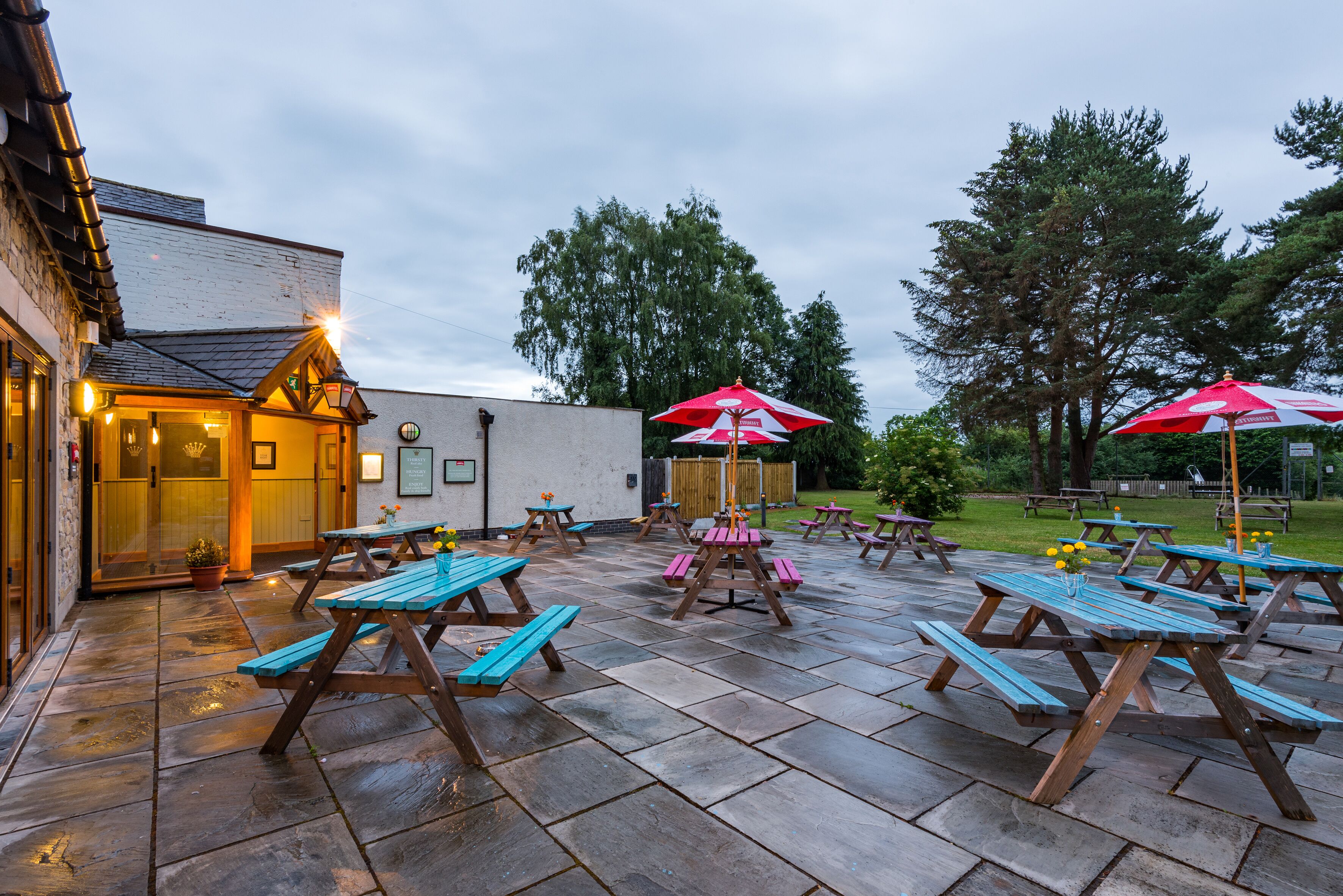A patio area with picnic tables and umbrellas, a building entrance, and a grassy area with trees.