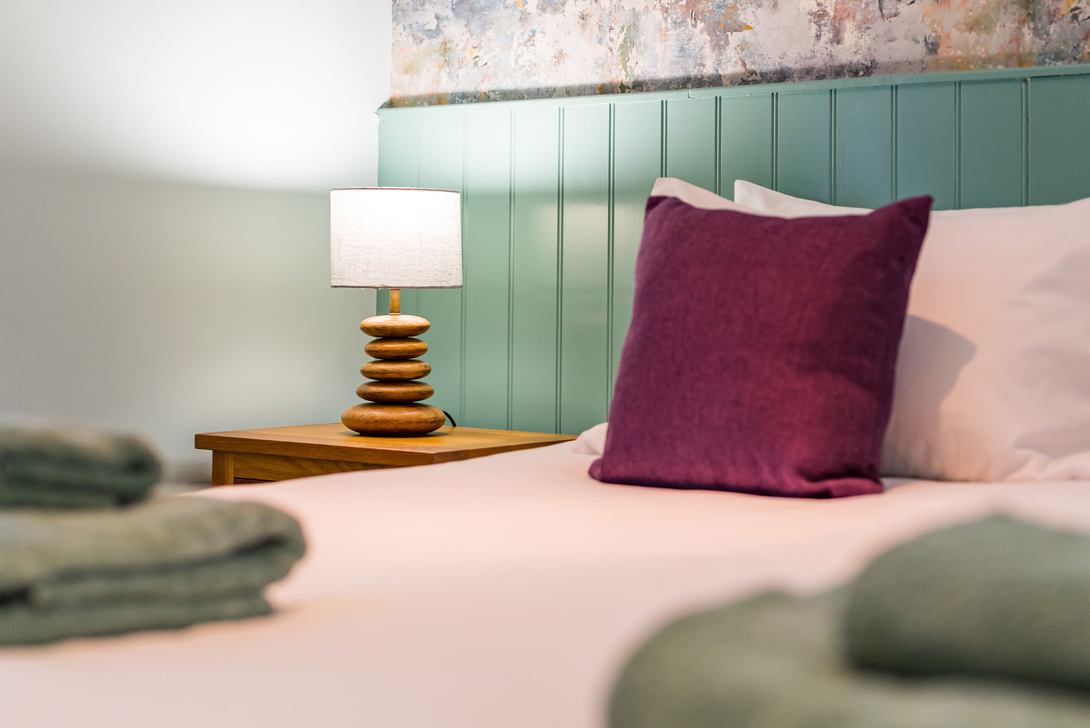 Een net opgemaakt bed met een houten nachtkastje ernaast, met een lamp en een stapel opgevouwen handdoeken.