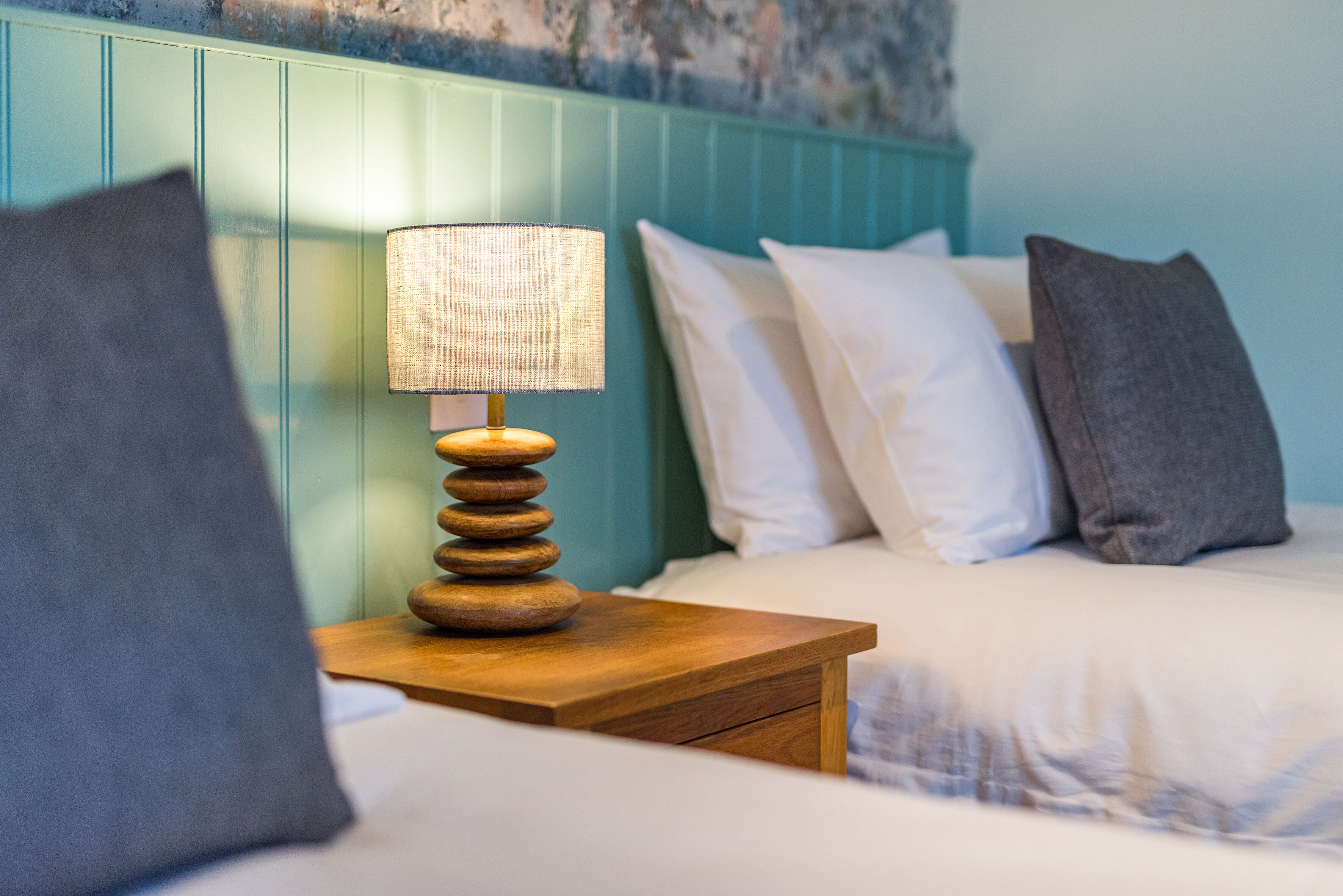 A neatly made bed with white and gray pillows, a wooden bedside table with a lamp, and a teal-colored headboard.