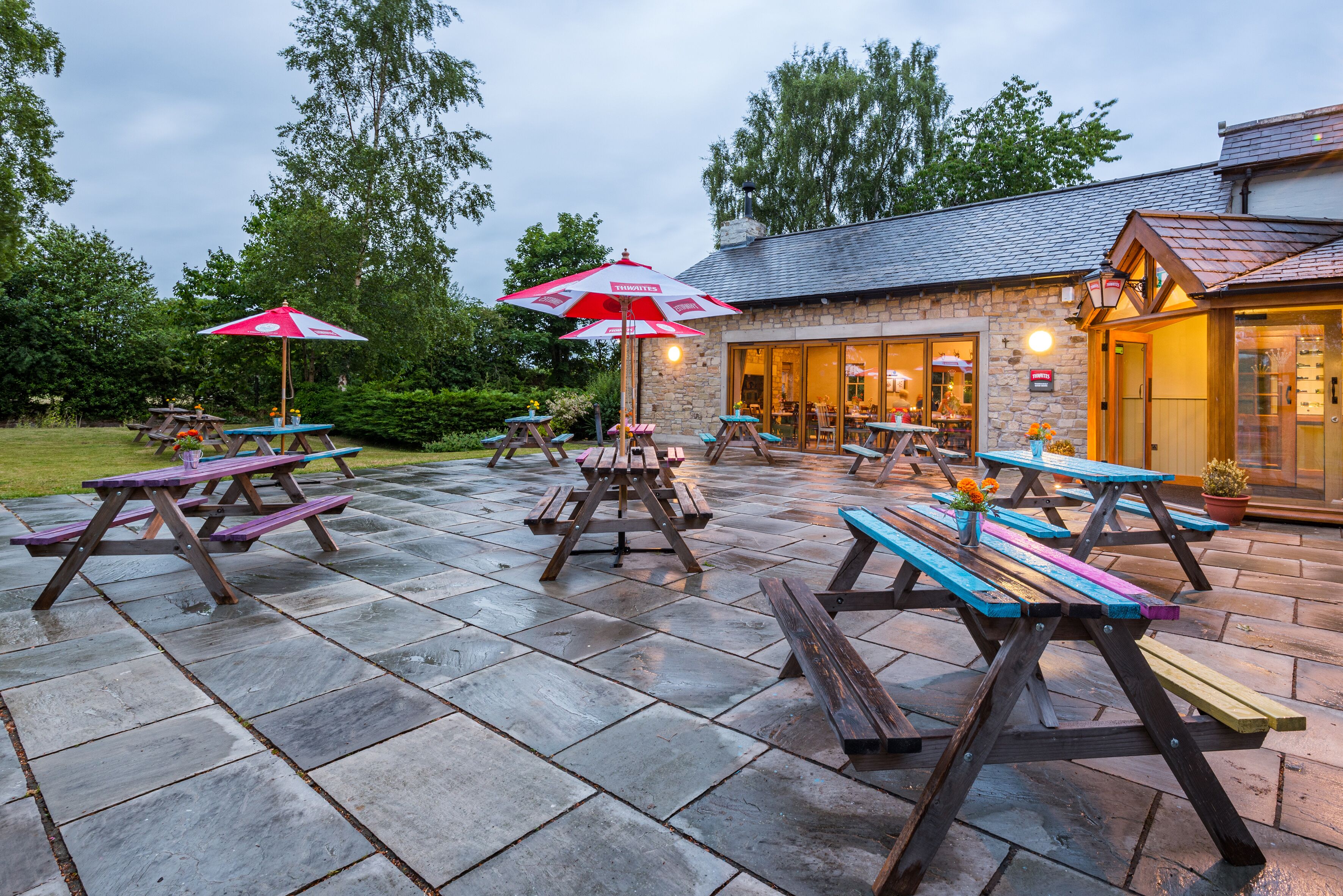 Een terras buiten met picknicktafels en parasols, stenen vloeren en een stenen gebouw met schoorsteen op de achtergrond.