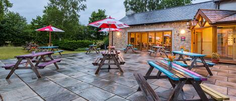 Een terras buiten met picknicktafels en parasols, stenen vloeren en een stenen gebouw met schoorsteen op de achtergrond.