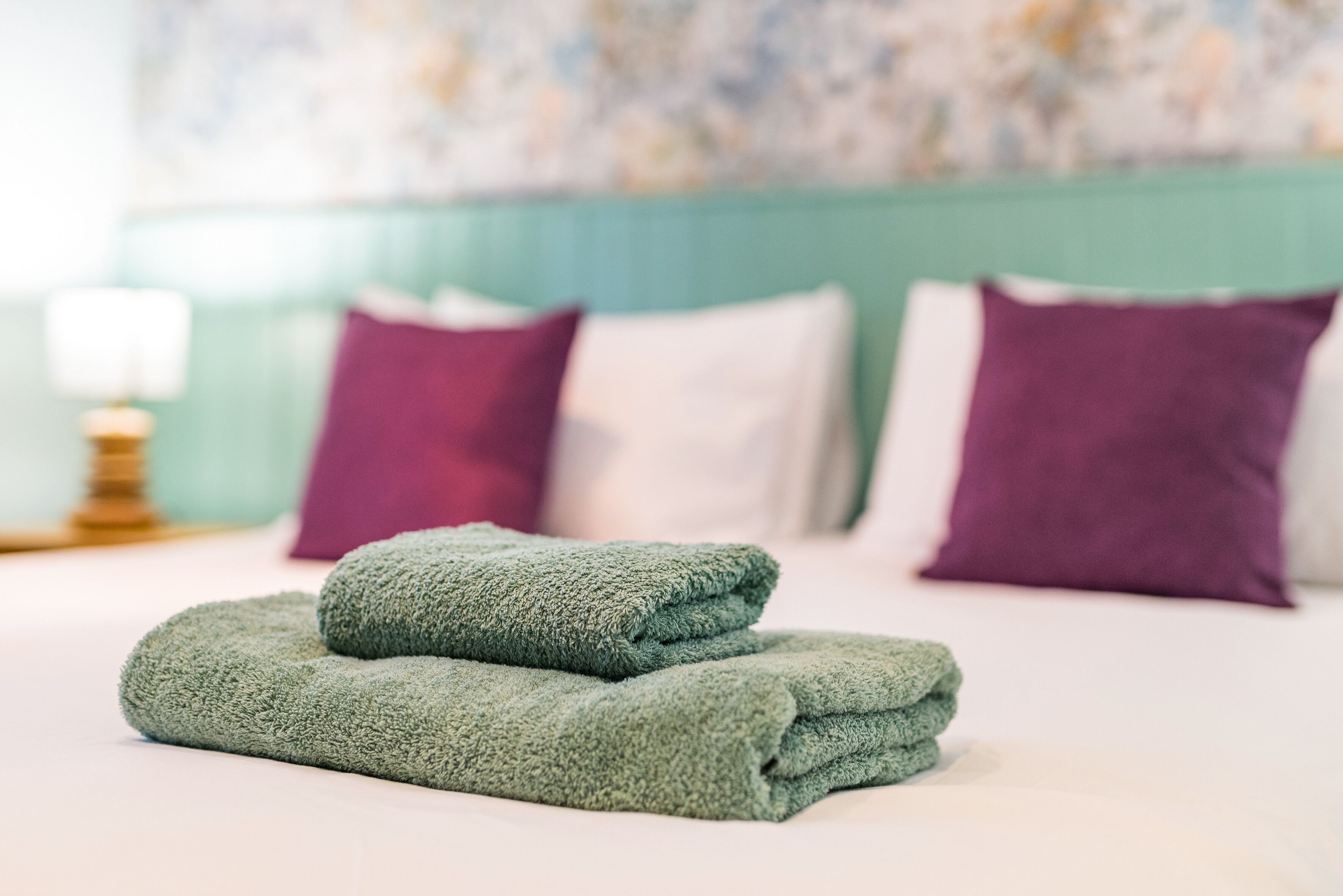 Een keurig opgemaakt bed met groene handdoeken, een blauwgroen hoofdeinde en paarse kussens.