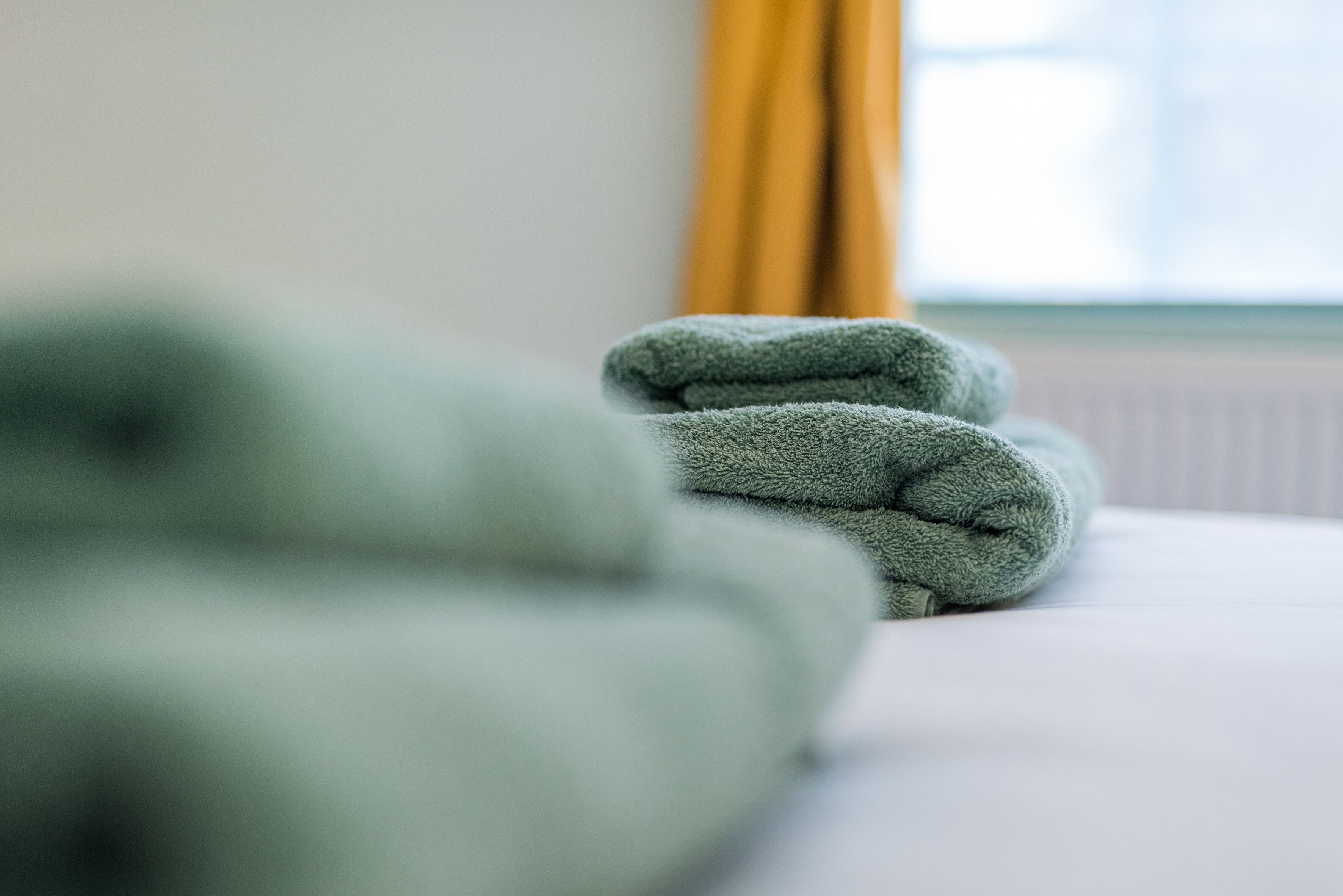 A neatly folded green towel on a white bed in a room with a window and yellow curtains.