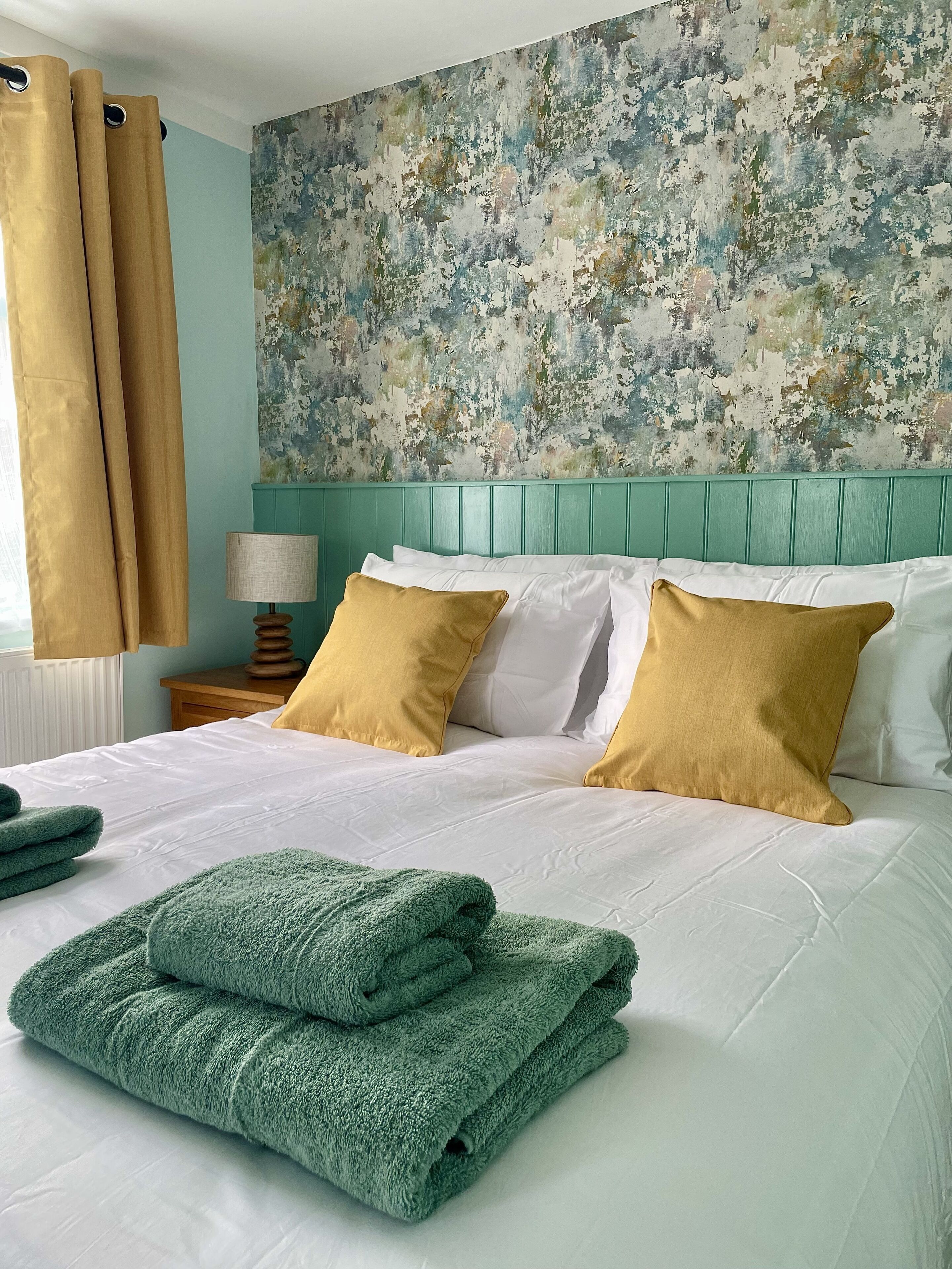 A neatly made bed with white and mustard pillows, green towels, and a teal headboard.