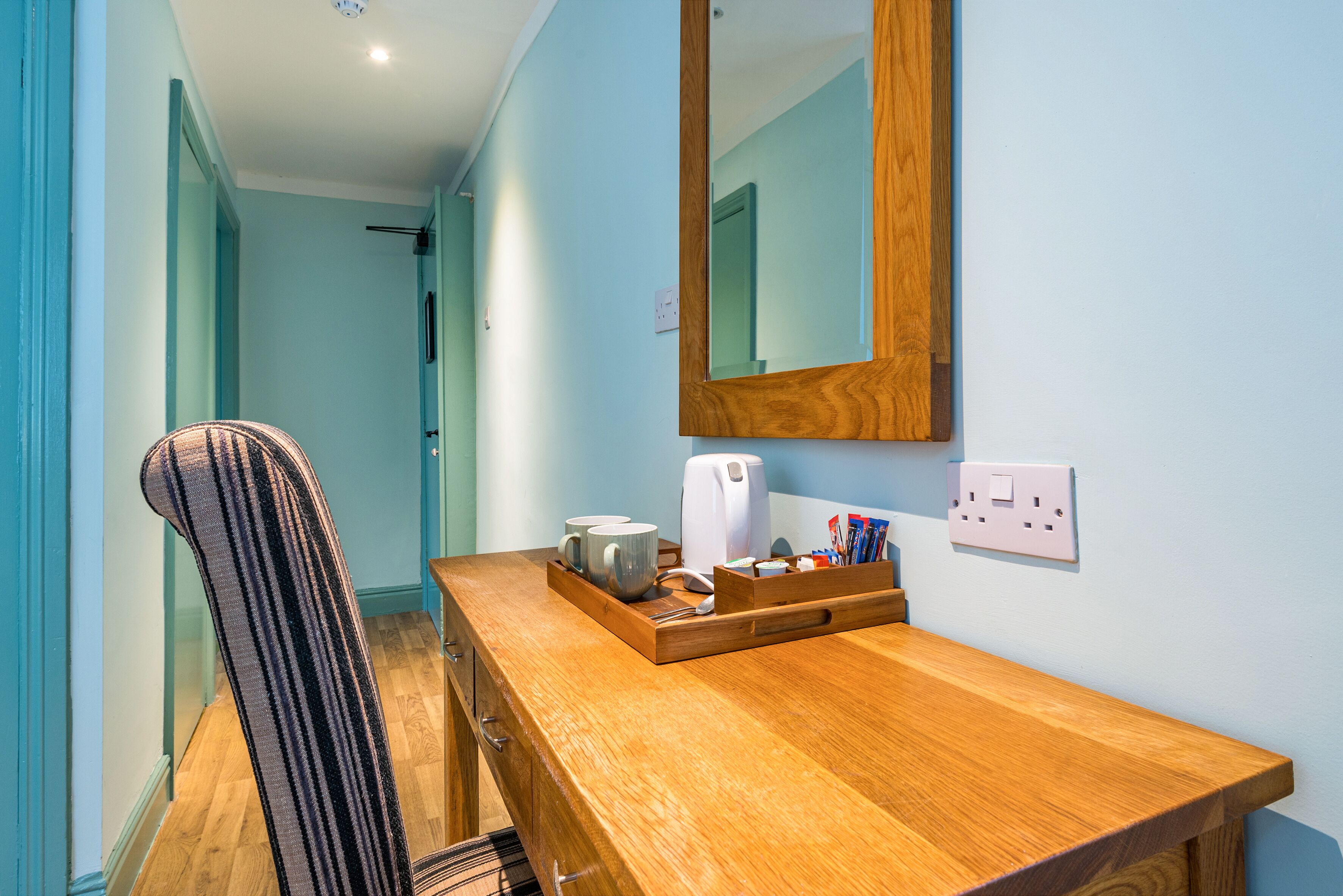 A wooden desk with a mirror, a tray with mugs, and a coffee maker in a room with light blue walls.