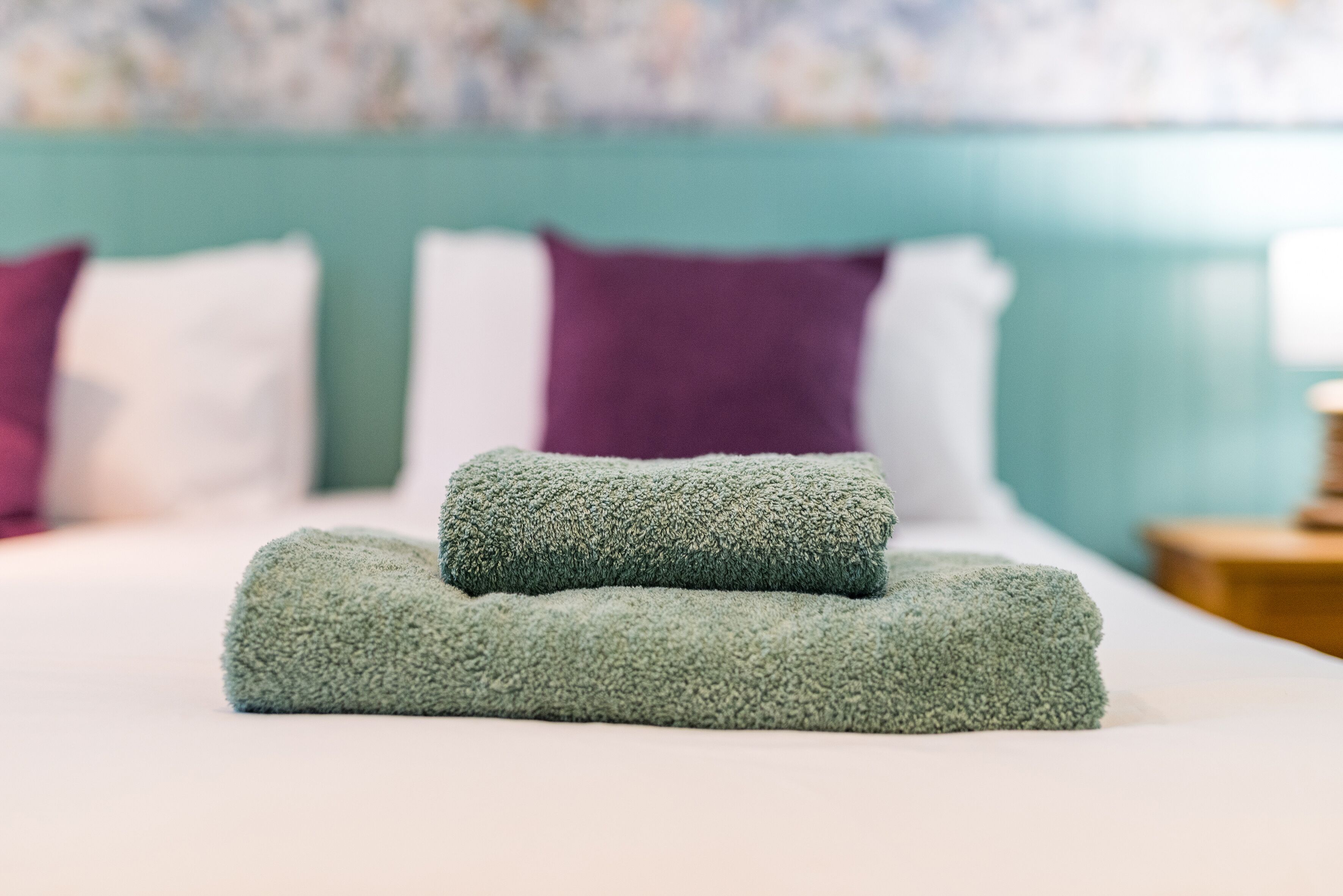 A neatly made bed with two folded green towels and a teal headboard.
