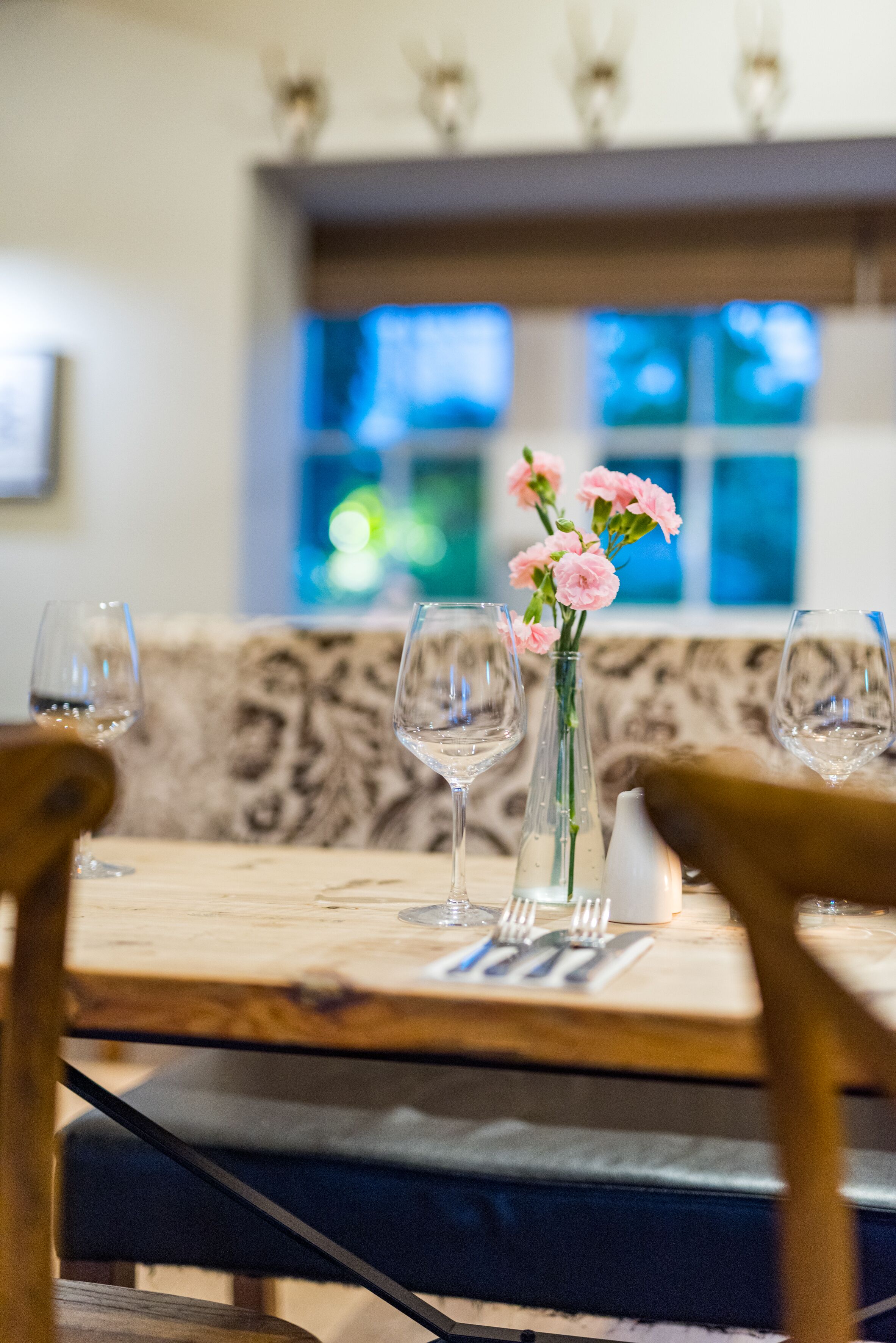 A dining table with a vase of pink flowers, wine glasses, and cutlery.
