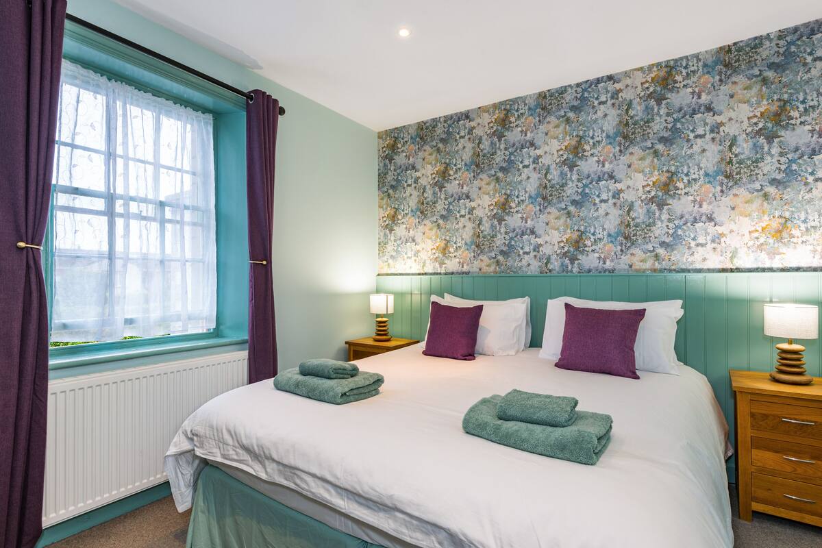 Un lit bien fait, agrémenté de serviettes vertes, une fenêtre avec des rideaux violets et un mur orné d’un motif floral.