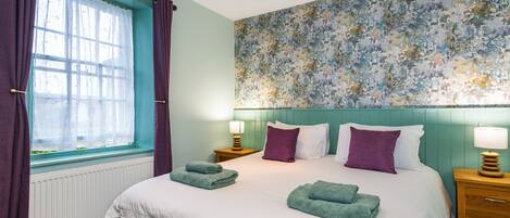 Una cama bien hecha con toallas verdes, una ventana con cortinas moradas y una pared con estampado floral.