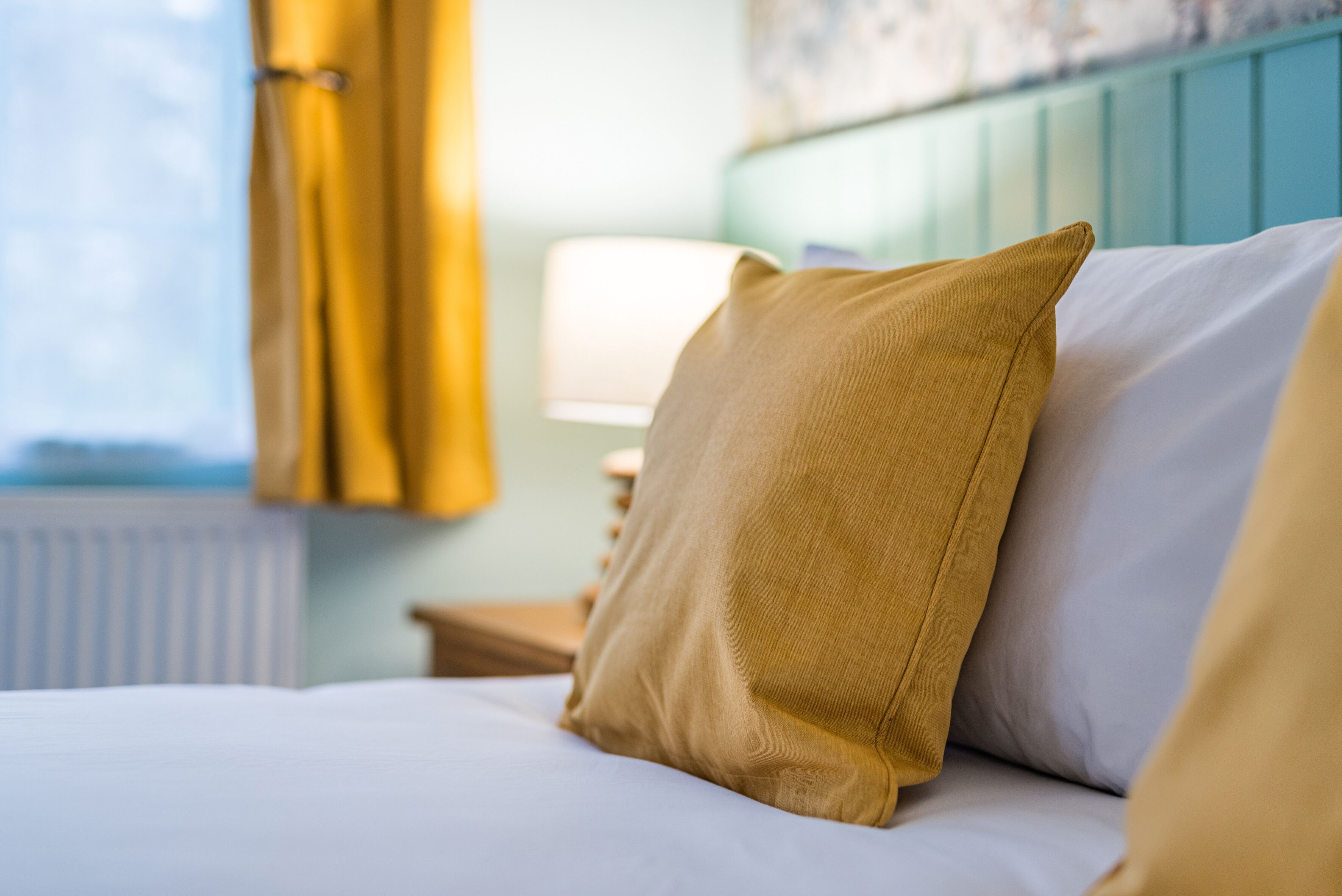 Una cama con un almohadón color mostaza y una almohadilla blanca, una mesita de noche con una lámpara, y una ventana con cortinas amarillas.