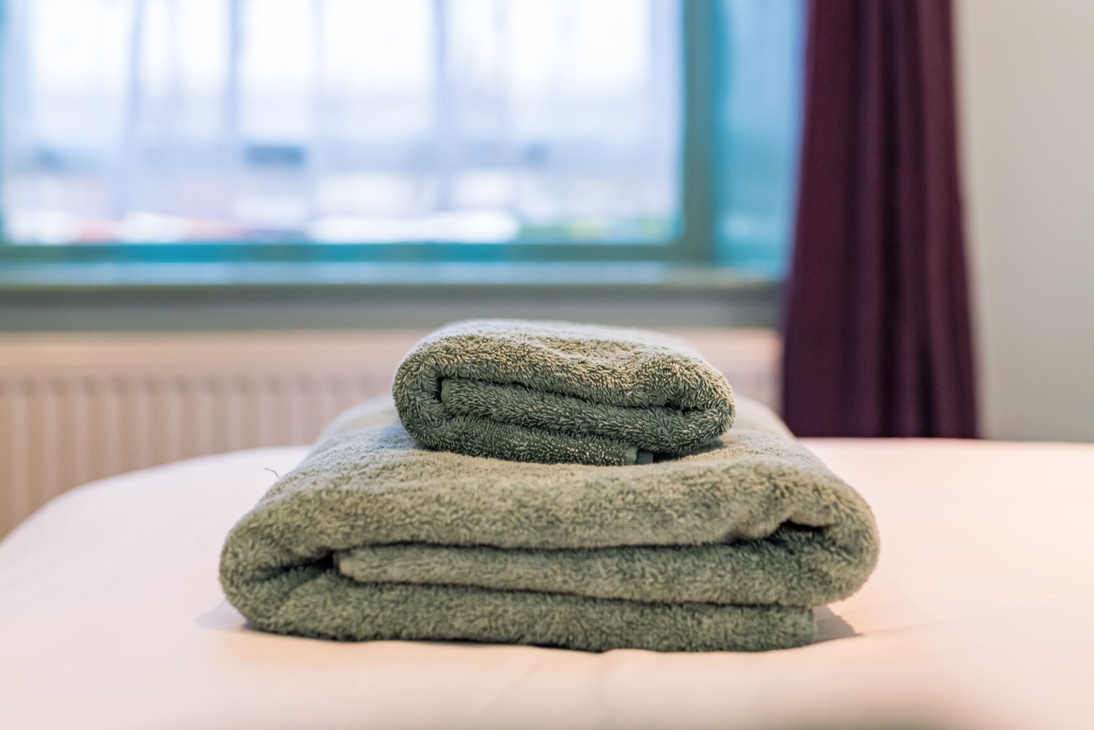 A neatly folded green towel stacked on a bed in a room with a window and a radiator.