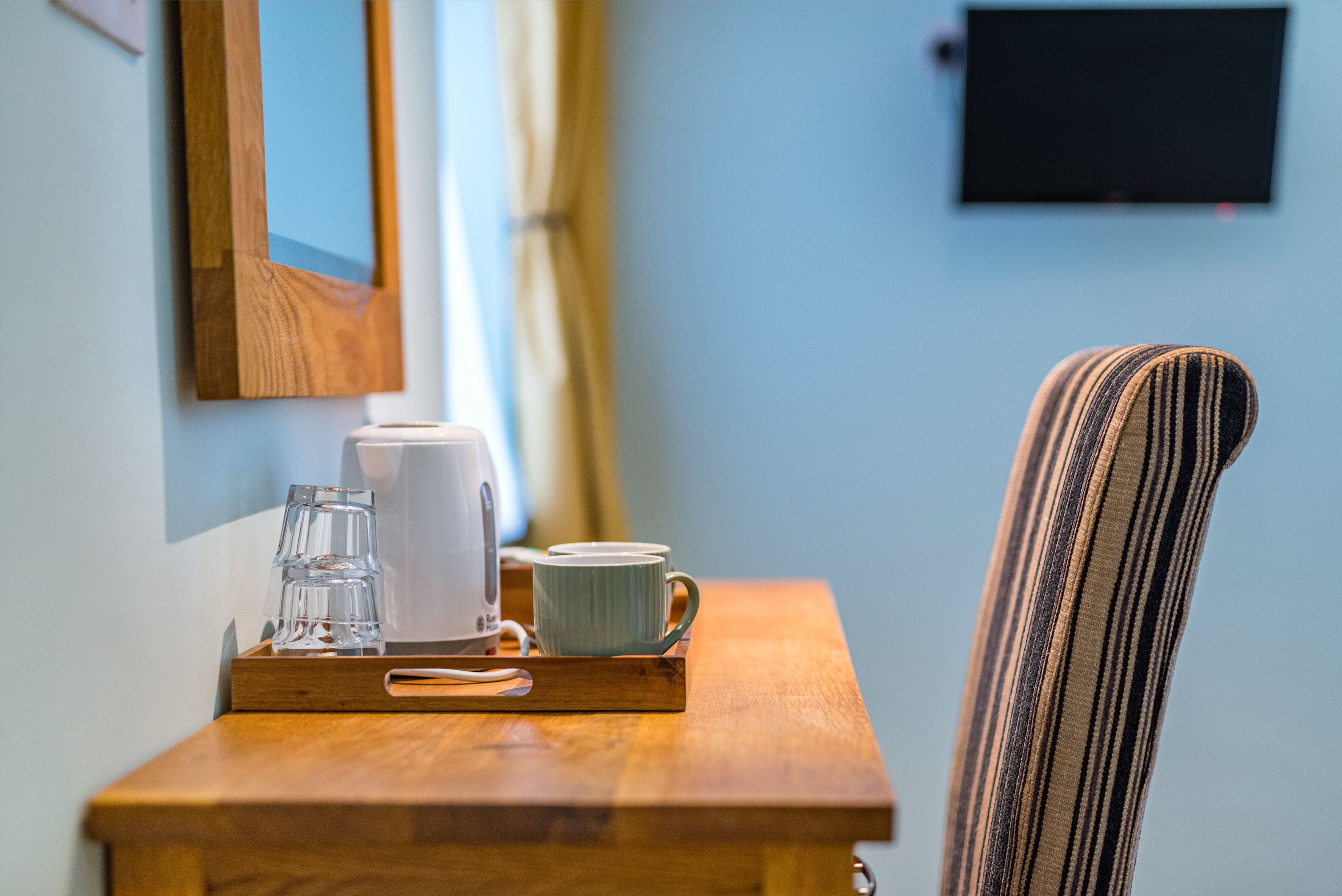 Una mesa de madera con una bandeja que sostiene un vaso, una taza y un hervidor. Una silla a rayas está a la derecha.