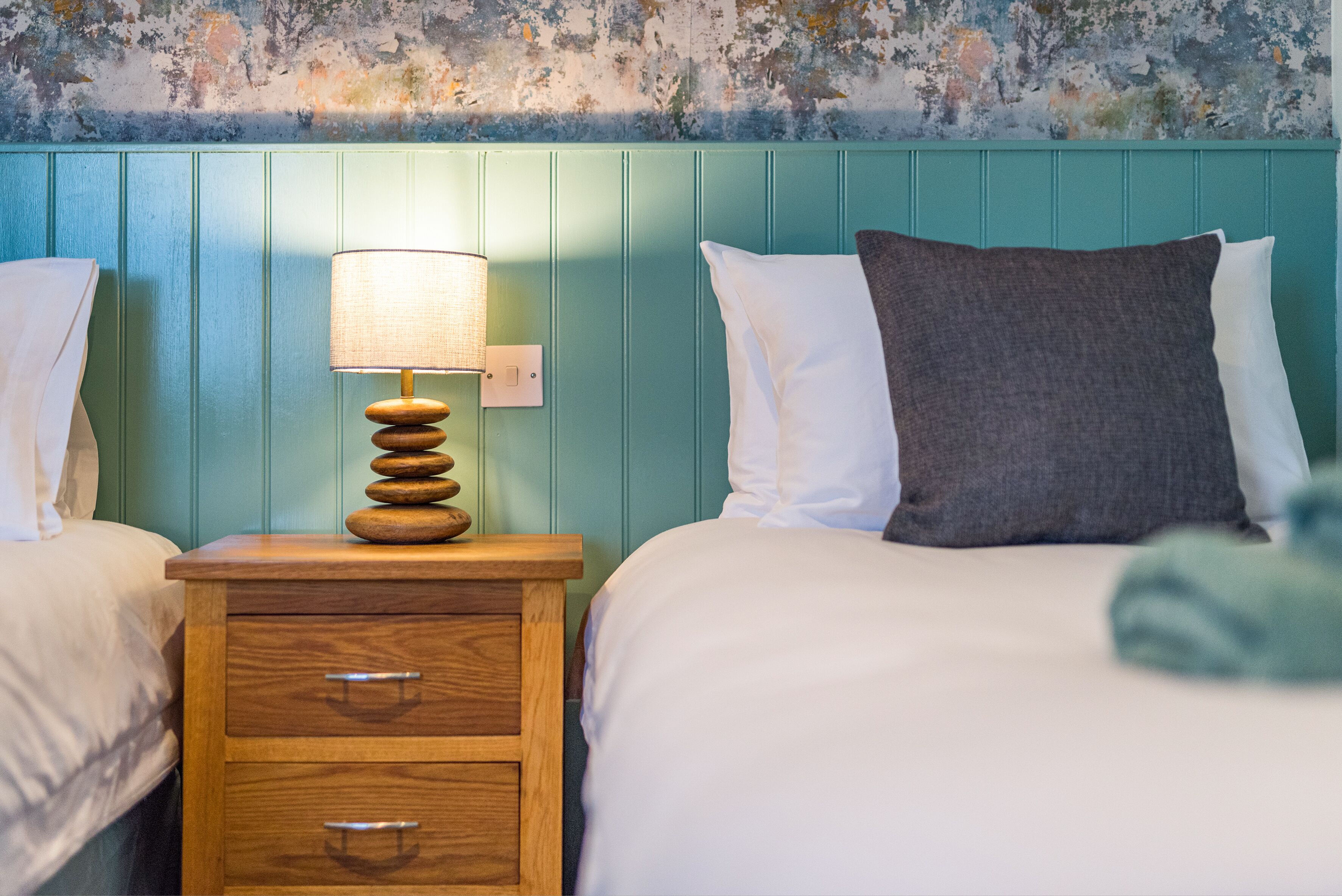 A bedroom with two beds, a wooden nightstand with a lamp, and a textured wall.