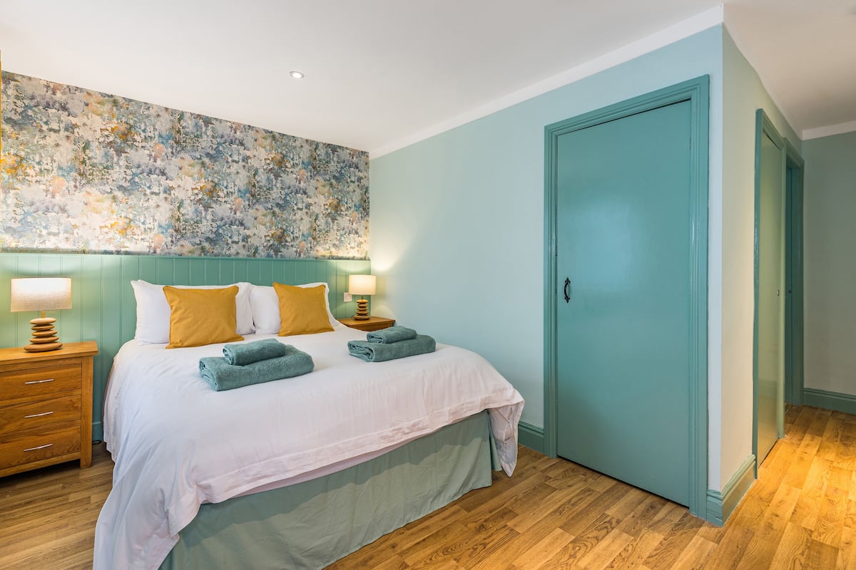Une chambre à coucher avec un lit, des tables de chevet, une porte de couleur turquoise et une fresque murale à motifs floraux.