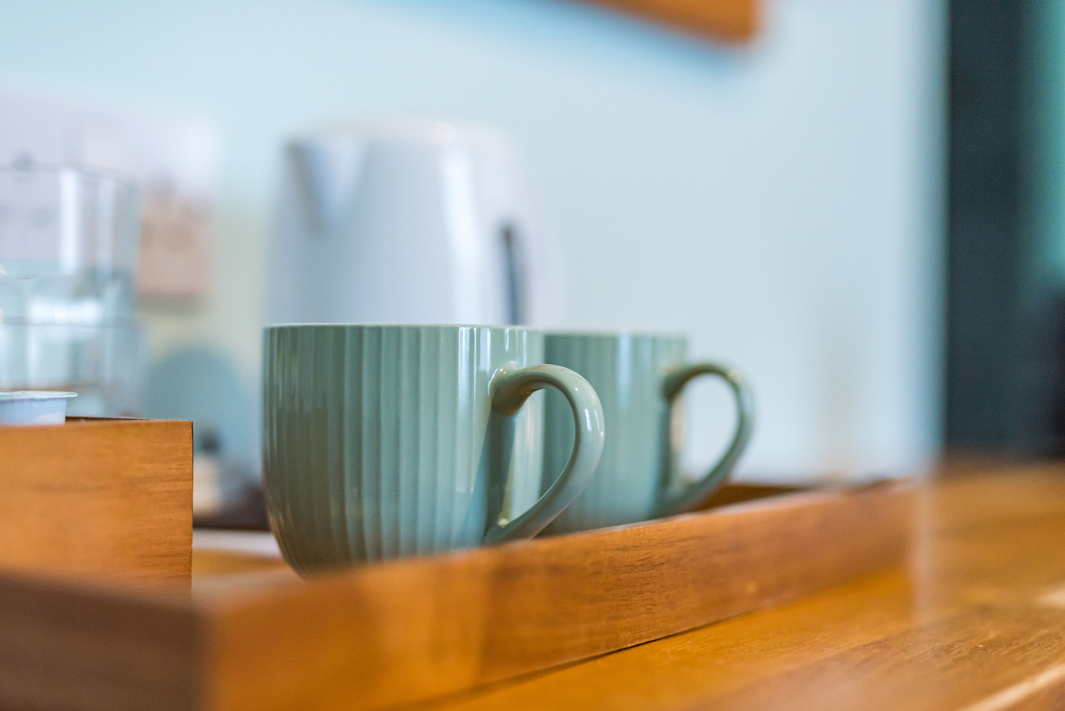 A wooden tray with two green ceramic mugs and a white electric kettle on a wooden table.