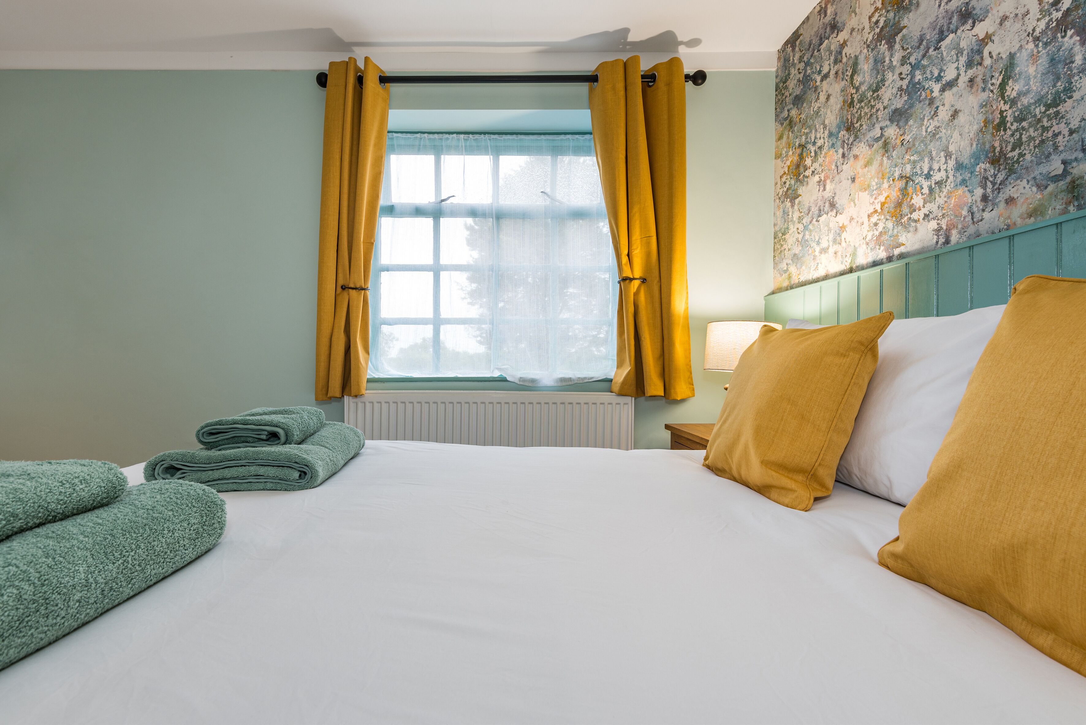 Een net opgemaakt bed met witte lakens, een raam met gele gordijnen en een groene handdoek ernaast.