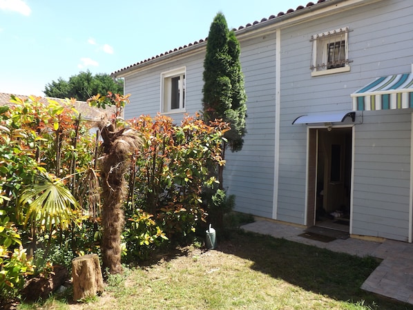 Una casa residencial con un pequeño jardín delantero, un árbol alto y un garaje con un toldo a rayas.