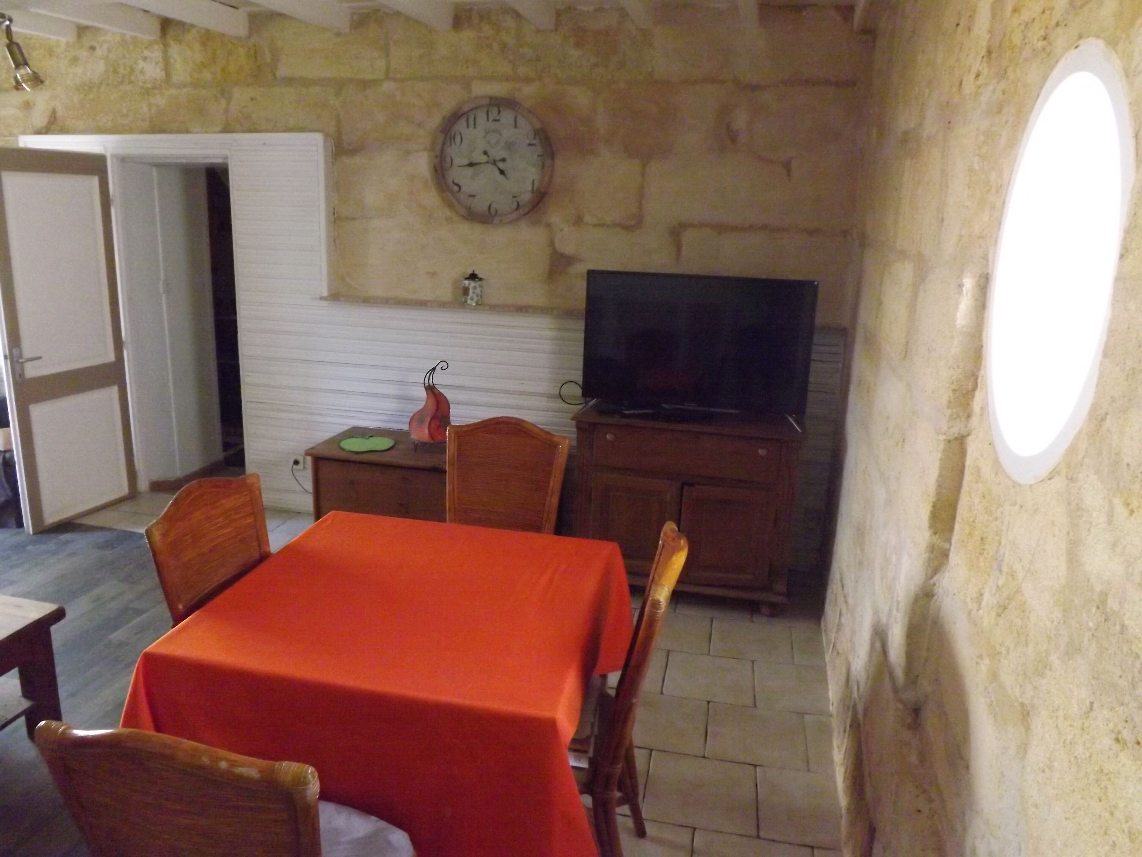 Uma sala de jantar com uma mesa posta para quatro pessoas, uma televisão sobre um móvel de madeira e um relógio na parede.