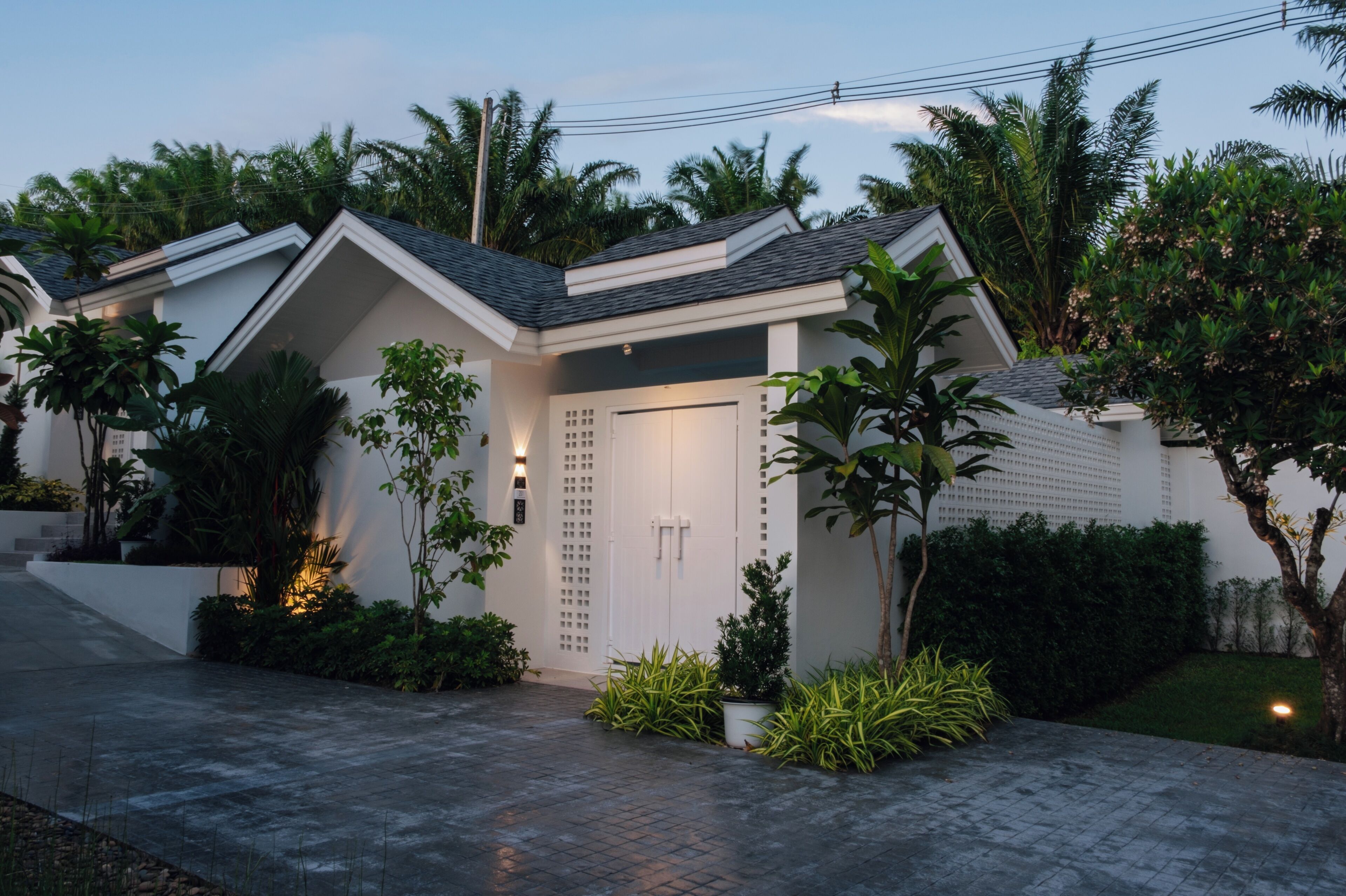 Une maison moderne à l’extérieur blanc, avec un toit foncé et un jardin bien entretenu, agrémenté de diverses plantes et doté d’une allée pavée.