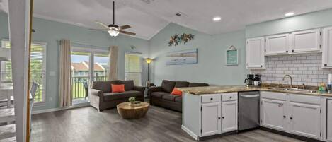 A spacious living area with a kitchen, featuring a ceiling fan, a dining table, and a sofa with cushions.