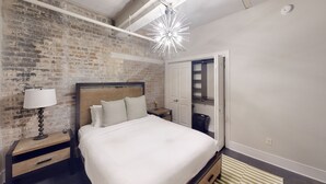 A bedroom with a brick wall, a modern chandelier, a bed with white bedding, a nightstand with a lamp, and a wardrobe with a mirror. - 60 Night Minimum Rental - FQ Apt. 2B/2B (New Orleans)