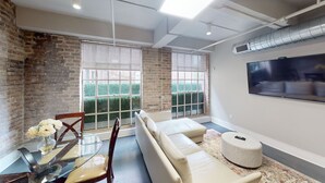 A modern living room with a brick wall, large windows, a sofa, a glass table, and a mounted TV. - 60 Night Minimum Rental - FQ Apt. 2B/2B (New Orleans)