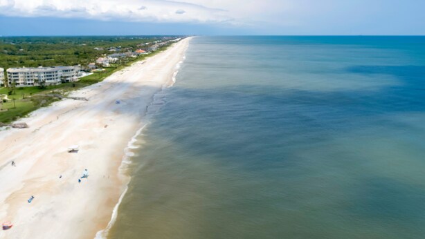 An aerial view of a beach with a long stretch of sand, a few buildings along the shore, and a clear sky. - Just steps to the beach and located within the Sawgrass Beach Club!  (Ponte Vedra Beach)