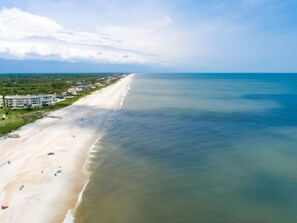 An aerial view of a beach with a long stretch of sand, a few buildings along the shore, and a clear sky.
