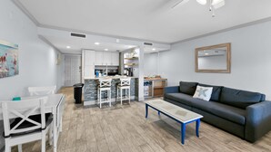 A modern living room with a dark grey sofa, a white dining table with chairs, a kitchen area with white cabinets, and a ceiling fan. - Just steps to the beach and located within the Sawgrass Beach Club!  (Ponte Vedra Beach)