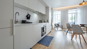 A modern kitchen with white cabinetry, a built-in oven, and a sink. There is a dining area with a table and chairs, and a large window providing natural light. - Nomad s Lux - Jardim Vista (Porto)