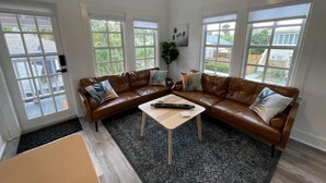 A living room with a brown leather sectional sofa, a small coffee table, and a patterned rug. There are large windows providing natural light and a view of the outside. - Modern 2BR Steps 2 Water Near Downtown! Fast Wifi (Lake Worth)