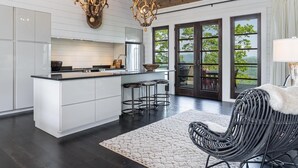 A modern kitchen with white cabinetry, a central island, and a dining area with a wicker chair and a view of the outdoors. - 5 Star Luxury, Lake Access, Spectacular Views! (Bryson City)