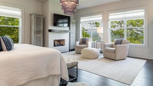 A spacious bedroom with a large bed, a fireplace, two armchairs, a round ottoman, and a TV mounted above the fireplace. - 5 Star Luxury, Lake Access, Spectacular Views! (Bryson City)