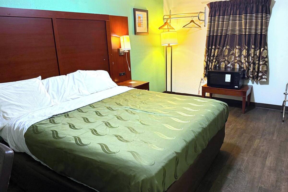 A hotel room with a bed, a nightstand, a lamp, a microwave, and a curtain.