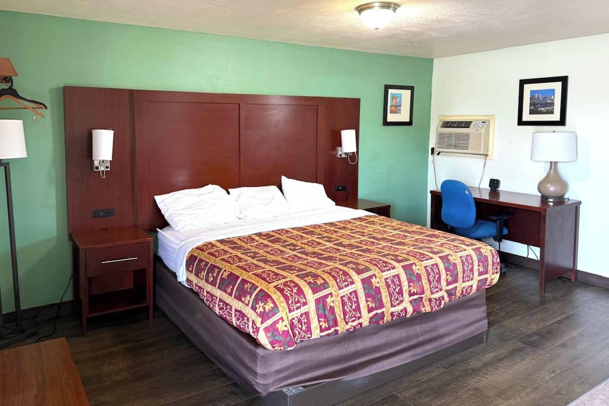 A hotel room with a large bed, a desk, a chair, a nightstand, and a wall-mounted air conditioner.