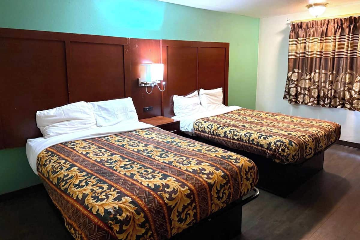 A hotel room with two beds, a nightstand, a wall-mounted lamp, and a window with curtains.