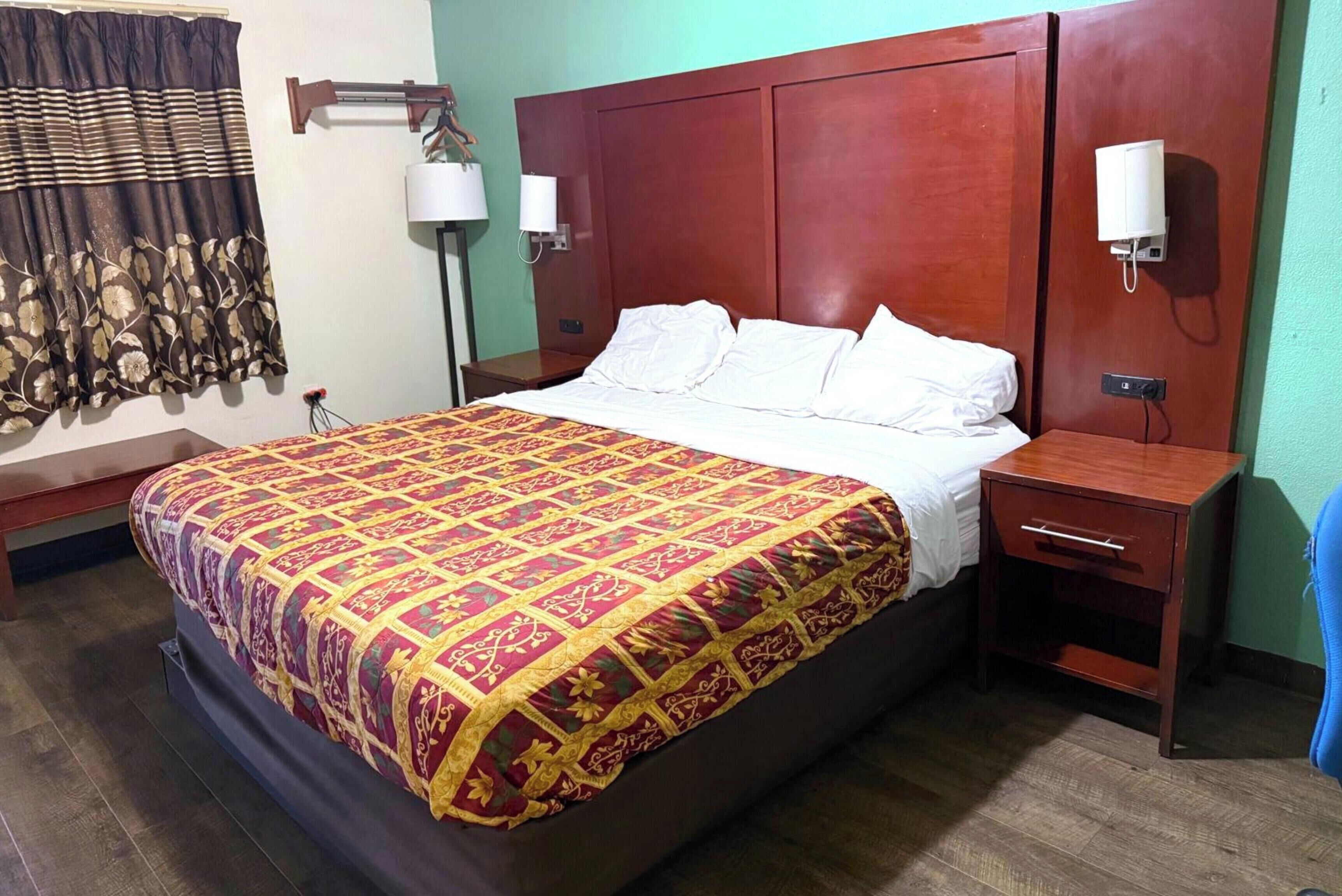 Een hotelkamer met een groot bed, een houten hoofdeinde, nachtlampjes, een nachtkastje en een raam met gordijnen.