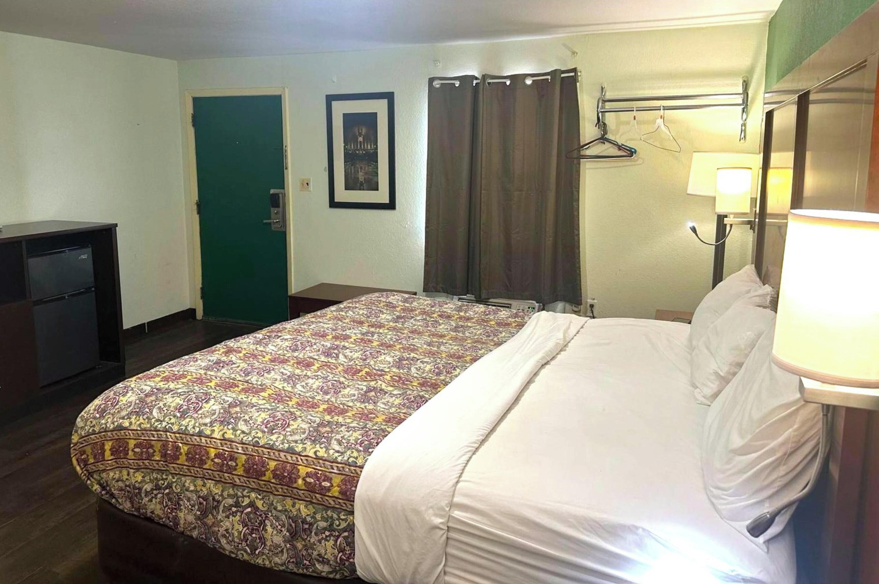 Ein Hotelzimmer mit einem Bett, einem Schreibtisch, einer grünen Tür, einem gerahmten Bild, einem Vorhang und einer Lampe.