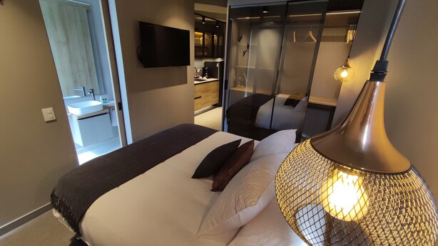 Camera d'hotel moderna con un letto, un comodino, una lampada, una televisione e un bagno parzialmente visibile attraverso una porta aperta.