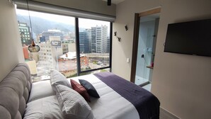 Una camera da letto con un'ampia finestra, un letto con molti cuscini, una TV a parete e vista sulla città.