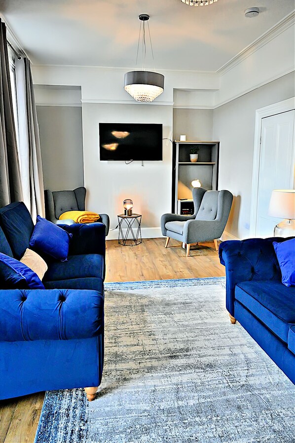 A modern living room with two blue sofas, a grey armchair, a small side table with a lamp, a flat-screen TV mounted on the wall, and a bookshelf.