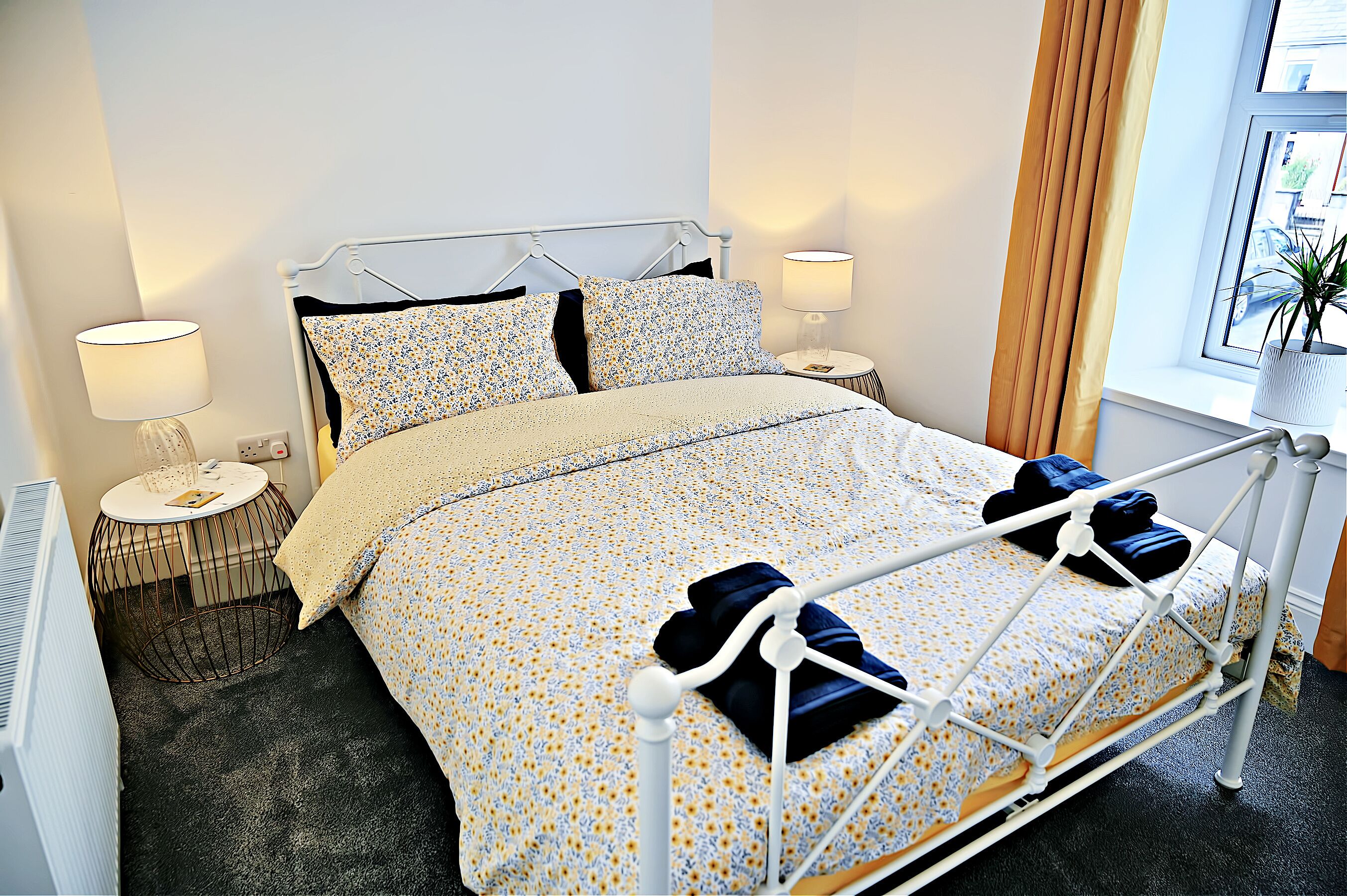A neatly made bed with a floral bedspread and two pillows, a bedside table with a lamp, and a window with curtains.