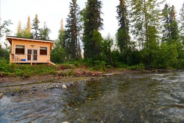 A wooden cabin by a river surrounded by tall trees.