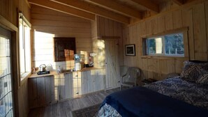 Interior de una cabaña de madera con una cama, una silla, una pequeña cocina y un ventanal.