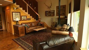 A spacious living room with a staircase, leather sofas, a patterned rug, and various wall decorations including antlers. - The Rusty Salmon Lake House with Deck and Dock (Talkeetna)