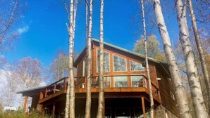 A wooden cabin with large windows and a balcony, surrounded by tall trees. - The Rusty Salmon Lake House with Deck and Dock (Talkeetna)